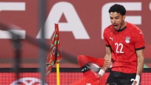 Un joueur égyptien célèbre joyeusement dans un kit rouge, avec un coéquipier au sol, entouré d'un drapeau marocain et d'un arrière-plan de stade vibrant.