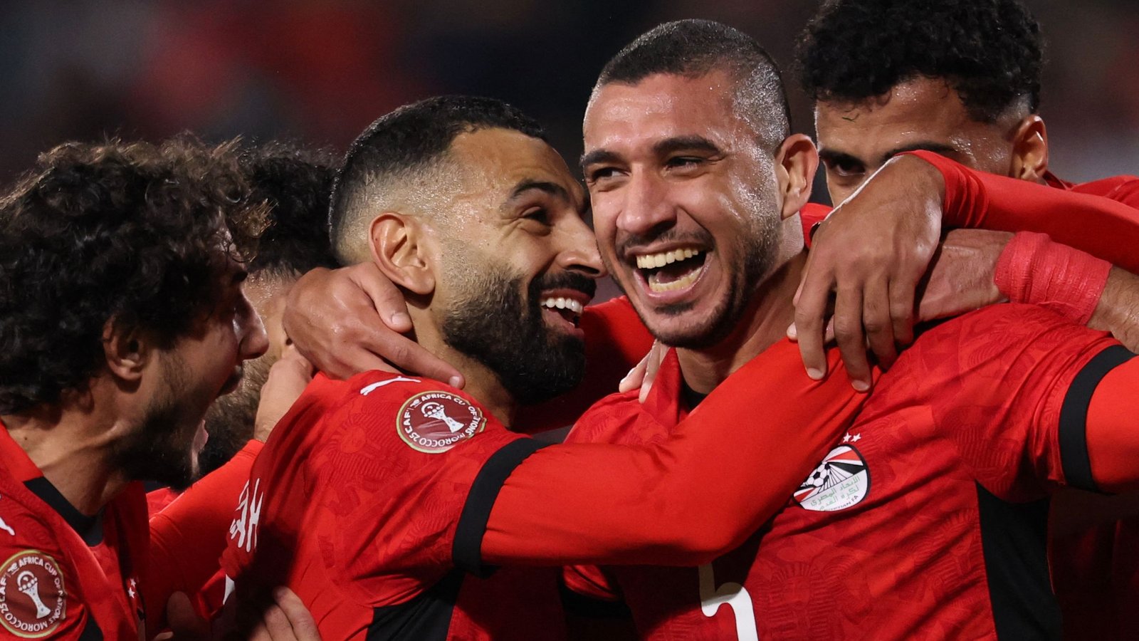 Les joueurs égyptiens célèbrent un but, portant des maillots rouges vifs, montrant joie et camaraderie pendant un match, avec des sourires et des étreintes partagées.
