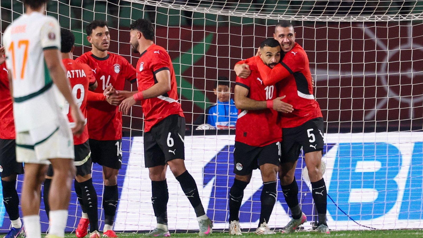 Egyptian players in red jerseys celebrate a goal, with a joyous embrace, during a match against an opposing team in white.