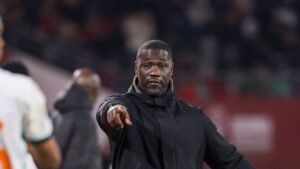 Coach in black jacket gestures passionately during a match, focused on guiding his team, with fans and players in the background.