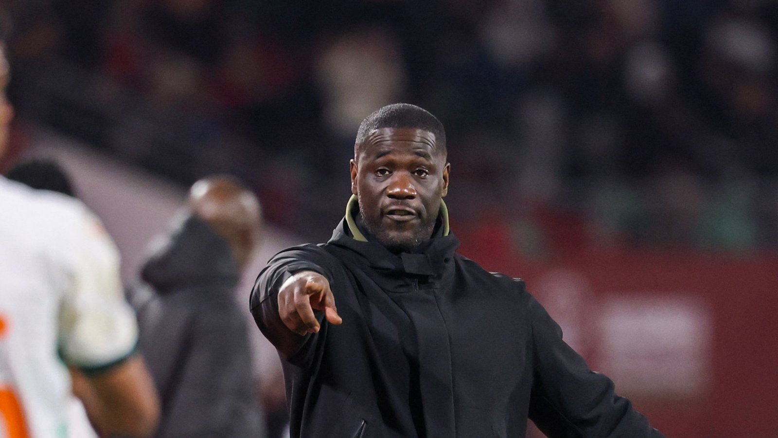 Coach in black jacket gestures passionately during a match, focused on guiding his team, with fans and players in the background.