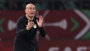 L'entraîneur de football marocain donne des instructions passionnées à son équipe pendant un match, portant un survêtement noir, avec un écusson de fédération en arrière-plan.