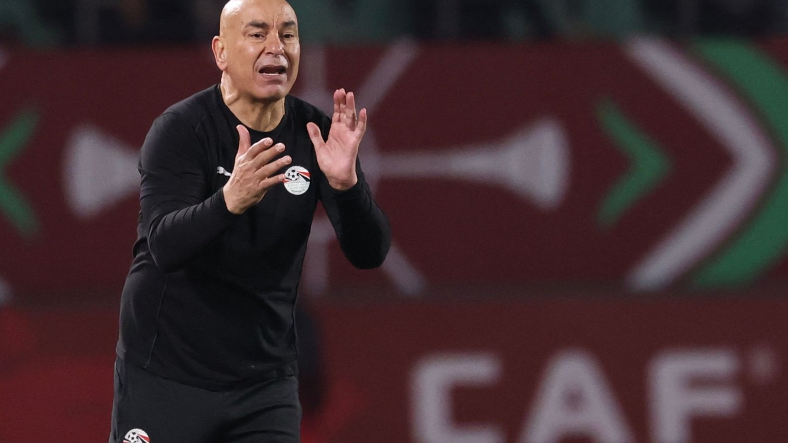 Entraîneur égyptien donnant des instructions avec passion aux joueurs lors d'un match de la CAF, portant un survêtement noir avec l'emblème de l'équipe nationale dans un stade vibrant.