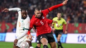 Ivory Coast's player in white challenges Egypt's defender in red during an intense match, with fans in the background cheering.