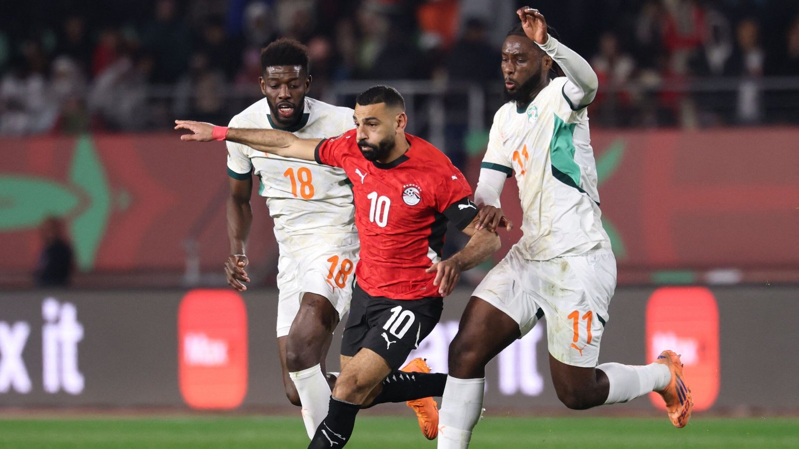 Mohamed Salah dans le maillot rouge de l'Égypte, dribblant des défenseurs ivoiriens lors d'un match intense de la CAN, capturé dans un cadre de stade.