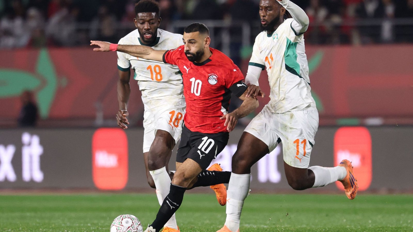 Mohamed Salah en tenue rouge d'Égypte, évitant deux défenseurs en blanc et vert, lors d'un match intense dans un stade plein.
