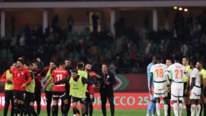 Les joueurs marocains célèbrent sur le terrain après un match, portant des maillots rouges, tandis que les adversaires en blanc se rassemblent pour une discussion avec les officiels.