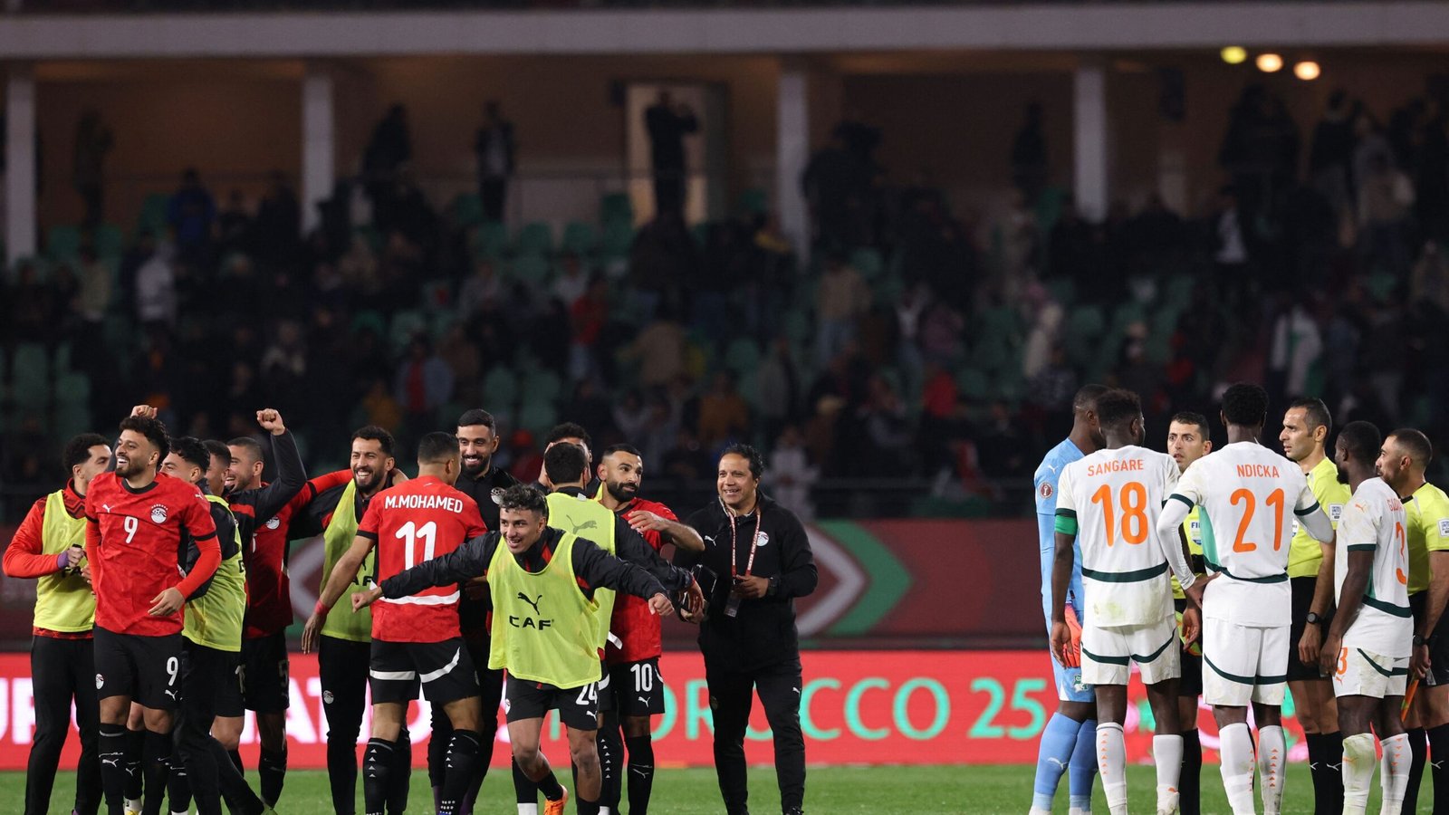 Les joueurs marocains célèbrent sur le terrain après un match, portant des maillots rouges, tandis que les adversaires en blanc se rassemblent pour une discussion avec les officiels.