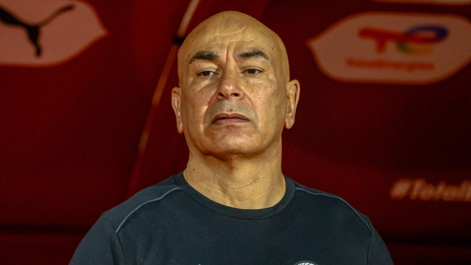 Moroccan football coach focused, wearing a black shirt, set against a red background featuring Puma branding.
