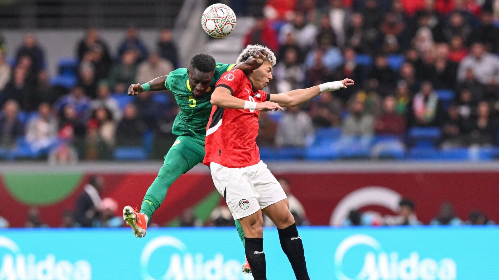 Senegal's player jumps to head the ball, challenging an Egyptian opponent during a tense moment in the match. Green and red team kits.