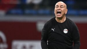 L'entraîneur égyptien dirige passionnément ses joueurs depuis la touche, portant un survêtement Puma noir avec l'écusson de l'équipe nationale, lors d'un match tendu.
