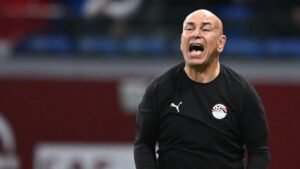 L'entraîneur égyptien criant avec passion pendant un match, portant un survêtement noir Puma avec le logo de l'équipe nationale, sur fond de stade flou.