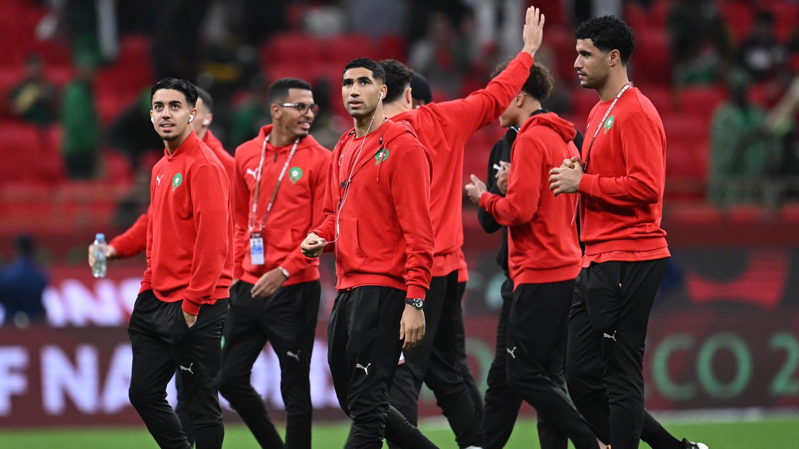 Les joueurs de l'équipe nationale marocaine, vêtus de tenues d'entraînement rouges, s'activent sur le terrain devant le fond vibrant d'un stade, avant un match.
