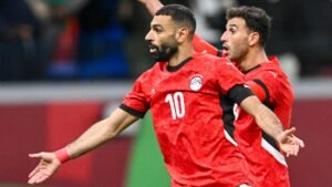 Mohamed Salah, in a vibrant red Egypt kit with number 10, celebrates a goal alongside a teammate in an intense match atmosphere.