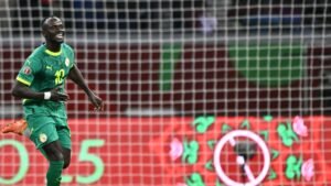 Sadio Mané celebrates in a green Senegal kit, scoring a goal during a match, with colourful background graphics and goal netting visible.