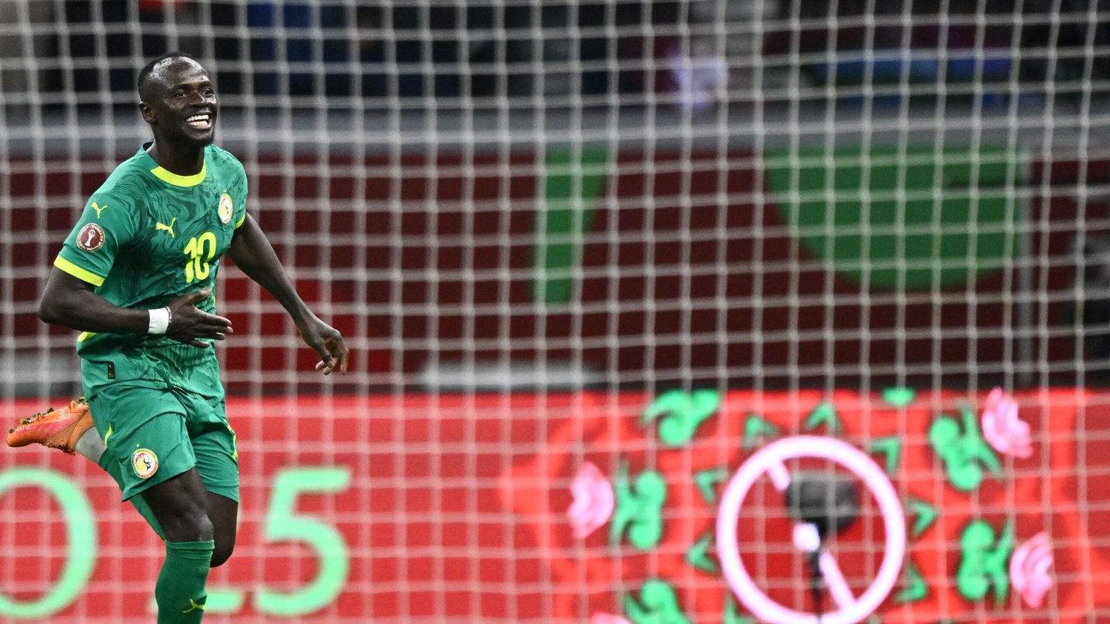 Sadio Mané celebrates in a green Senegal kit, scoring a goal during a match, with colourful background graphics and goal netting visible.