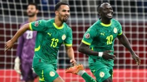 Sadio Mané and his teammate celebrate a goal, wearing vibrant green Senegal kits with yellow accents, in front of a net.