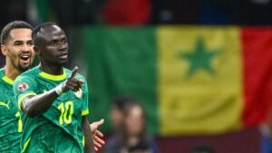 Sadio Mané célèbre avec un coéquipier, pointant la foule du doigt, tandis que le drapeau sénégalais flotte en arrière-plan, capturant l'esprit de l'équipe.