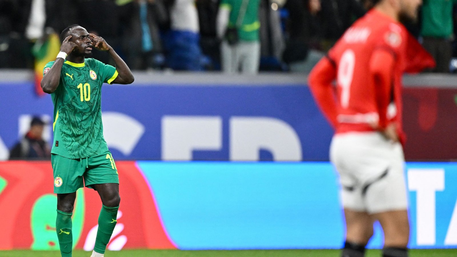 Sadio Mané, vêtu du maillot vert du Sénégal, exprime sa frustration par un geste lors d'un match, un adversaire en rouge apparaissant flou en arrière-plan, illustrant l'atmosphère compétitive.