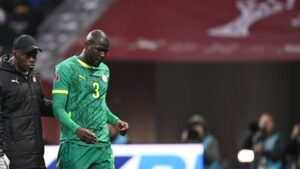 Senegalese player wearing a green kit with yellow accents walks off the pitch, assisted by a staff member during a match.