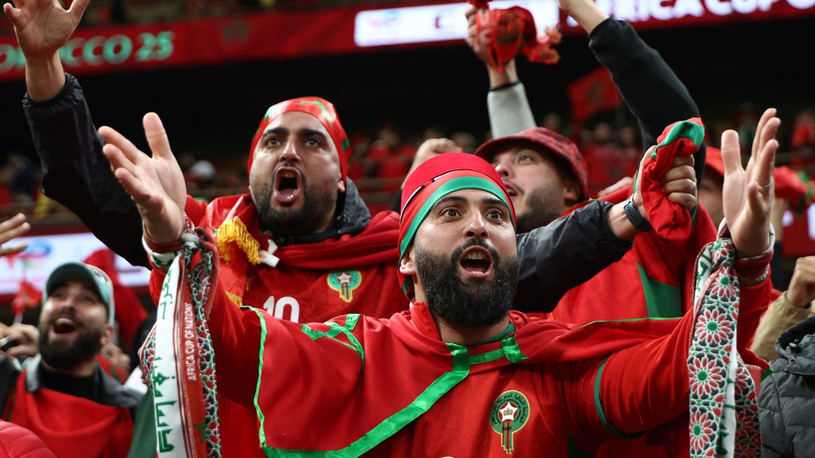 Moroccan fans passionately celebrating in vibrant red and green attire during an Africa Cup of Nations match, showcasing national pride and excitement.