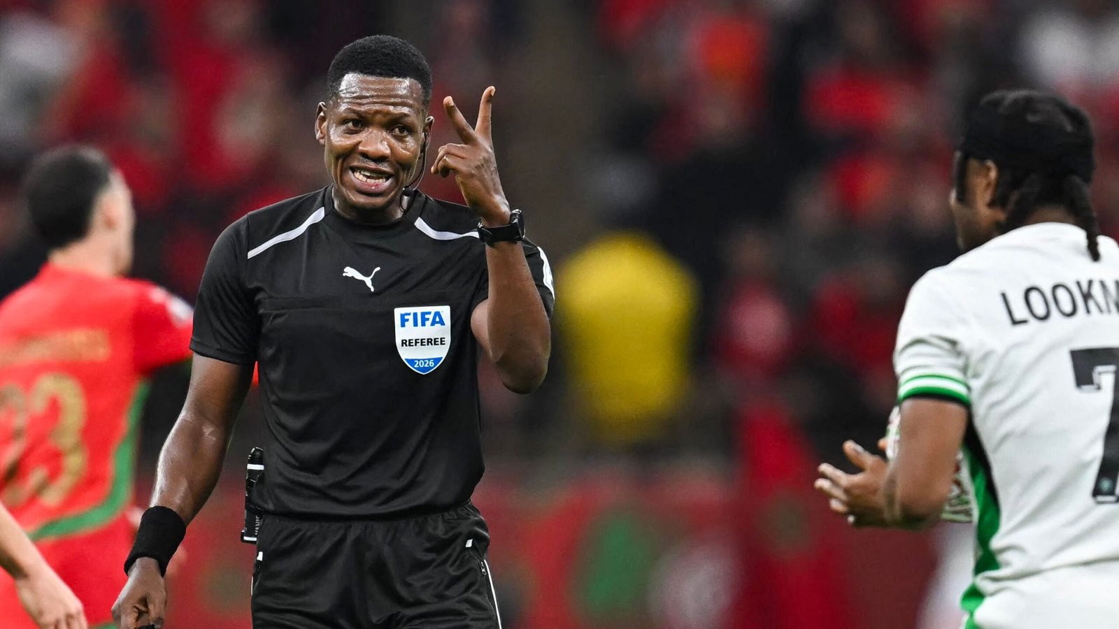 FIFA referee gestures during a match, engaging with a player wearing a green and white kit, against a vibrant stadium backdrop.