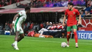 Moroccan player in red kit dribbles the ball past a Nigerian opponent in green during a tense match, with fans and players in the background.