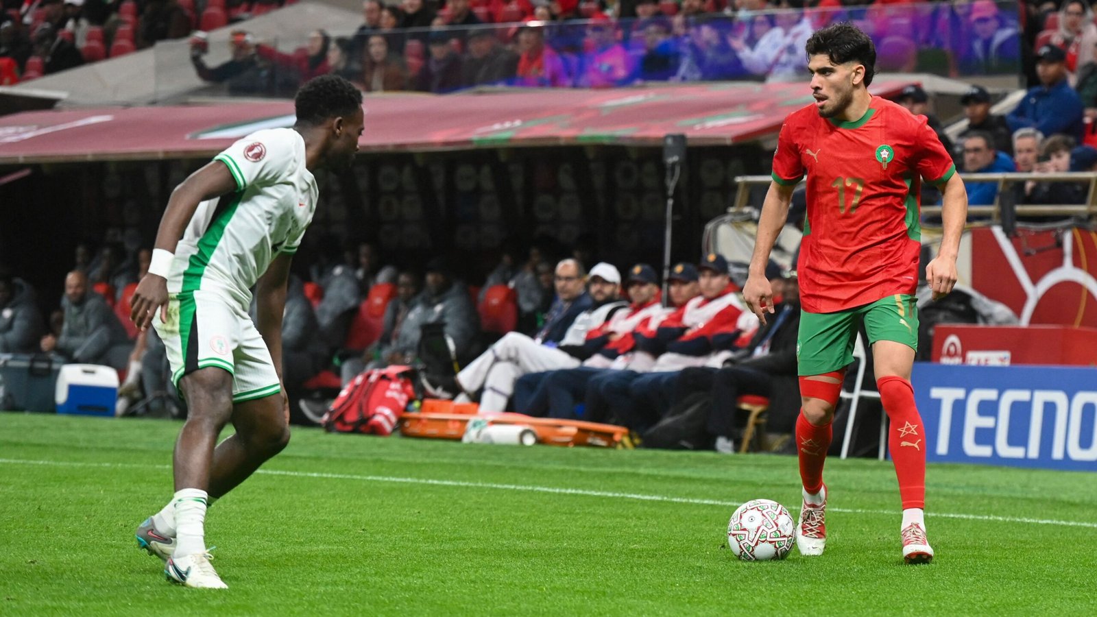 Moroccan player in red kit dribbles the ball past a Nigerian opponent in green during a tense match, with fans and players in the background.