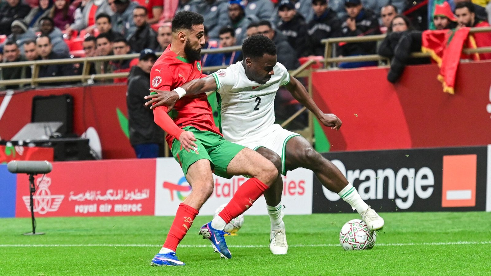 Un joueur marocain entre en confrontation avec un adversaire dans un moment dynamique durant un match, mettant en valeur le maillot rouge et vert vibrant de l'équipe.