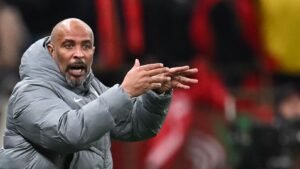 L'entraîneur de football marocain gesticule avec passion en bord de terrain, vêtu d'une veste grise, avec les supporters et les couleurs du stade flous en arrière-plan.