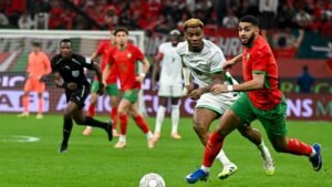 Les joueurs marocains pourchassent un adversaire lors d'un match tendu, arborant des tenues d'un rouge et vert vifs devant une foule animée.