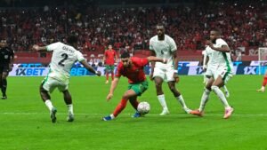 L'attaquant marocain tente de dribbler deux défenseurs dans un stade vibrant, avec des supporters passionnés en rouge visibles en arrière-plan.