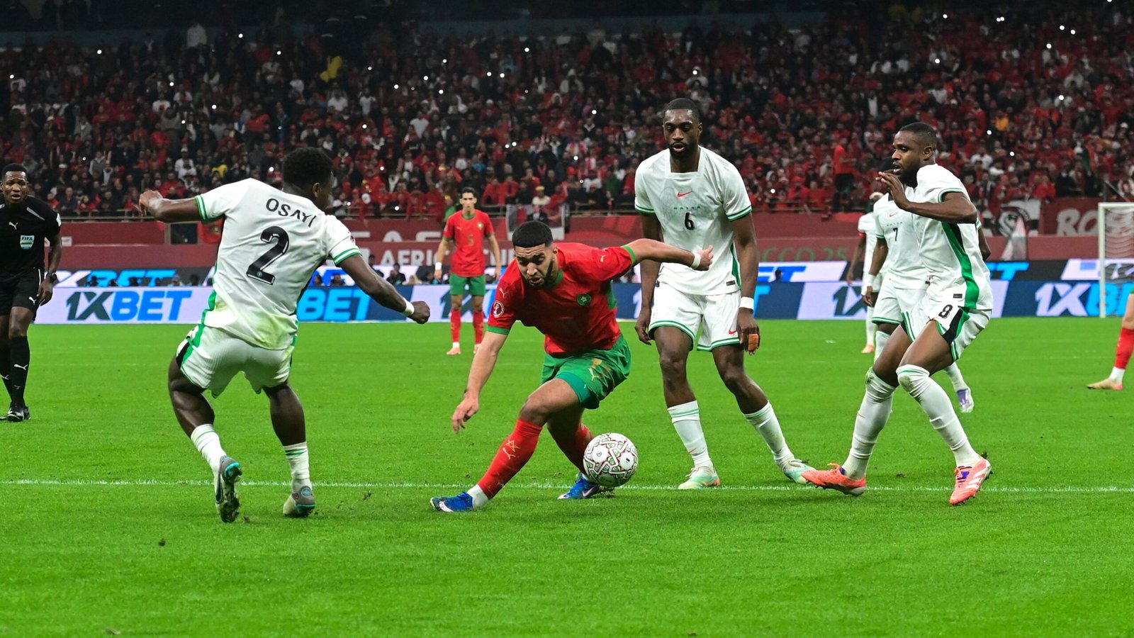 L'attaquant marocain tente de dribbler deux défenseurs dans un stade vibrant, avec des supporters passionnés en rouge visibles en arrière-plan.
