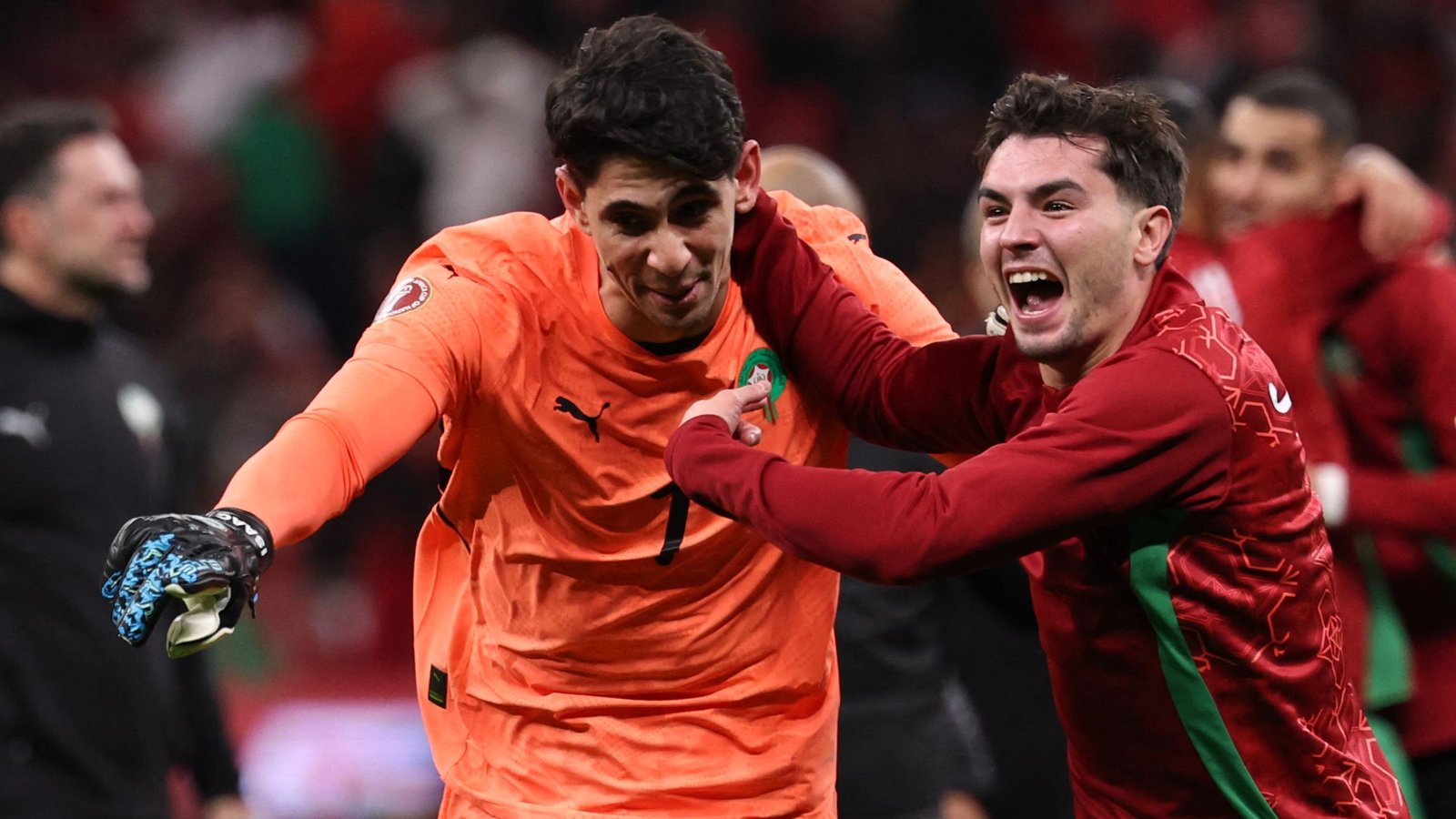 Moroccan players celebrate a victory, featuring a goalkeeper in an orange kit and a teammate in a red jersey, expressing joy.