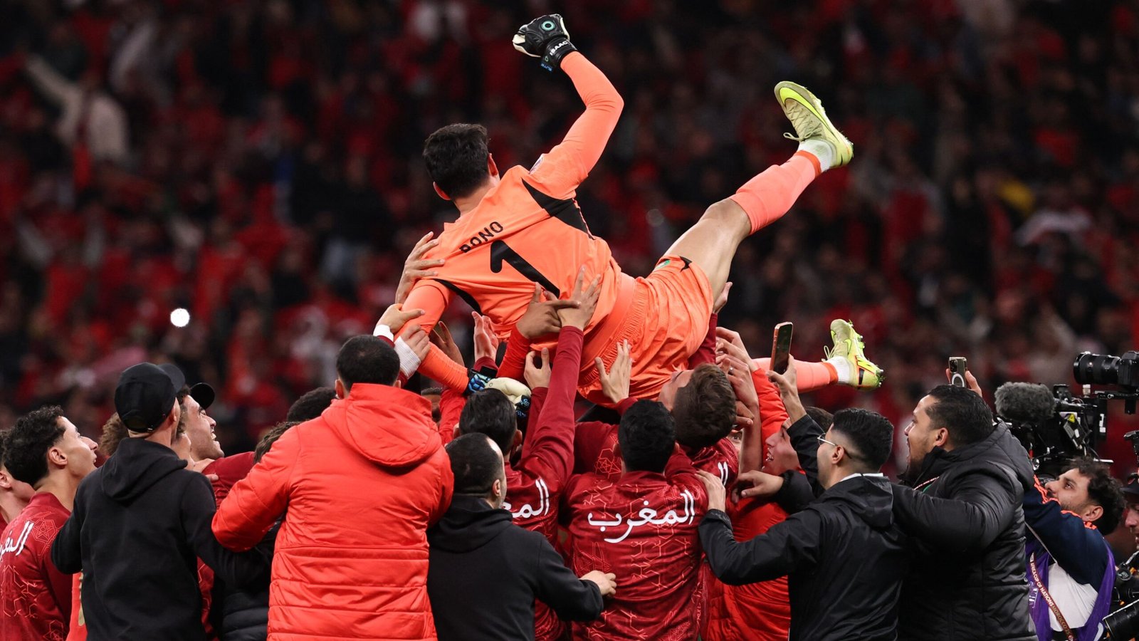 Bono, the Moroccan goalkeeper, is lifted joyfully by teammates in red jerseys, celebrating a significant victory amid a sea of cheering fans.