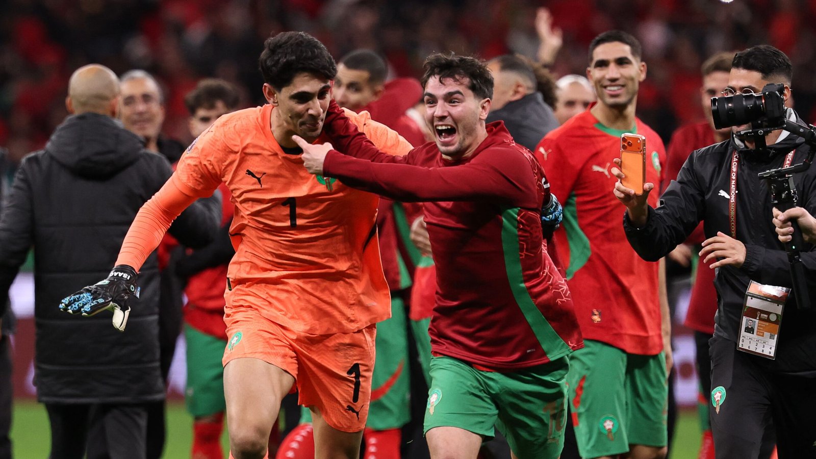 Moroccan players celebrate a victory, with goalkeeper Yassine Bounou in an orange kit and a teammate in red, amidst a jubilant crowd.