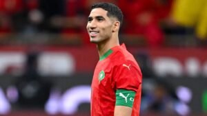 Achraf Hakimi smiles while wearing Morocco's red kit with green accents, showcasing his captain's armband during a match.