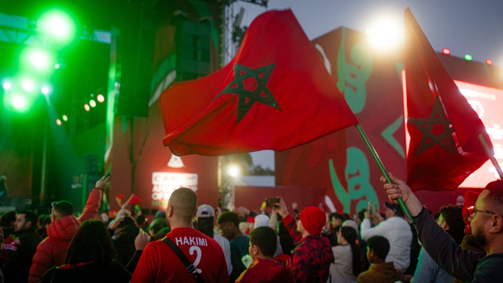 Les supporters marocains agitent des drapeaux rouges arborant l'emblème national, en liesse pendant le tournoi de la CAF, avec des projecteurs de scène colorés en arrière-plan.