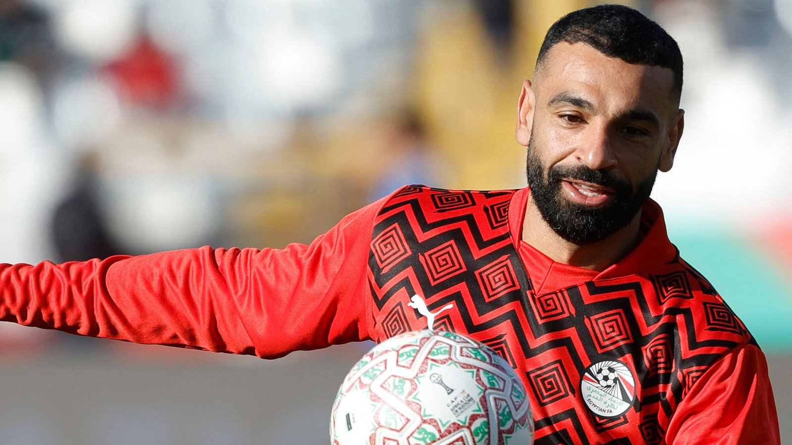 Mohamed Salah smiles while warming up in a vibrant red training kit with geometric patterns, holding an ornate football.