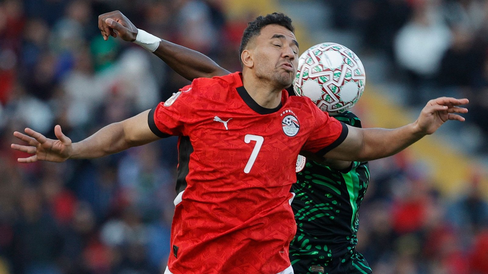 لاعب مصري يرتدي زيًا أحمر يتصادم مع خصم يرتدي اللون الأخضر، بينما تضرب كرة القدم وجهه خلال لحظة مشحونة في المباراة.