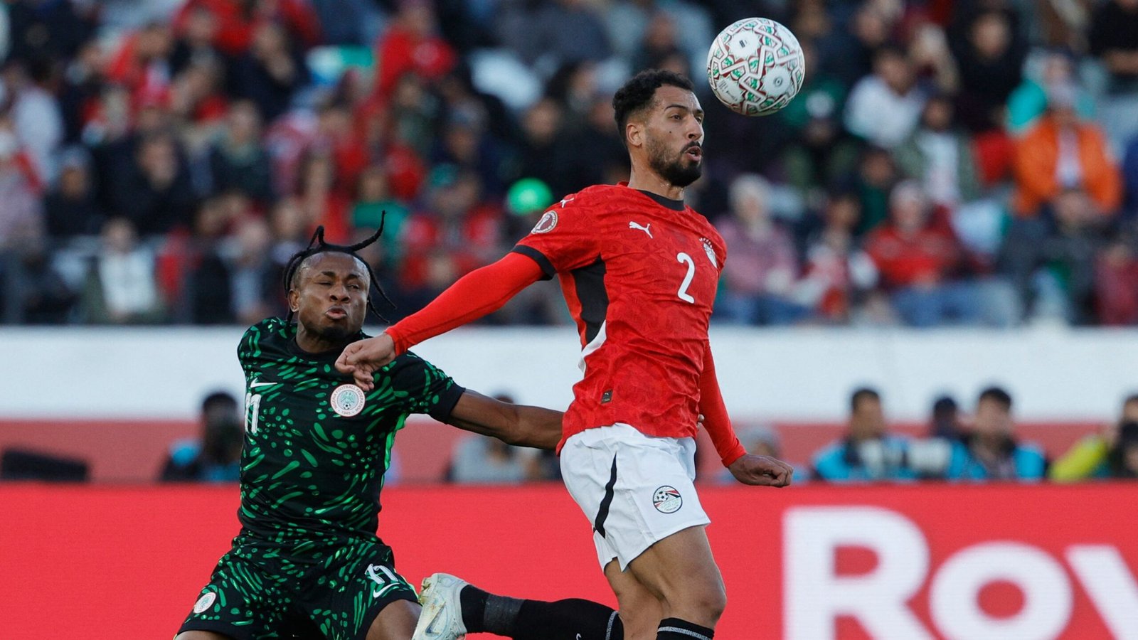 Egyptian defender in red challenges Nigerian player in green for the ball during an intense match. Fans watch in the background.