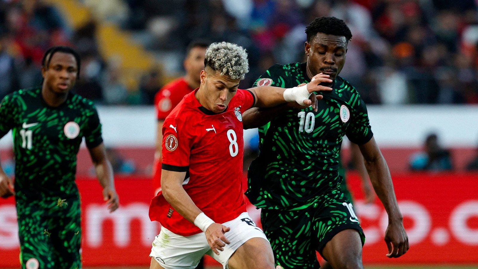 Moroccan player in red jersey No. 8 challenges Nigerian opponent in a dynamic moment during a competitive match, with fans in the background.