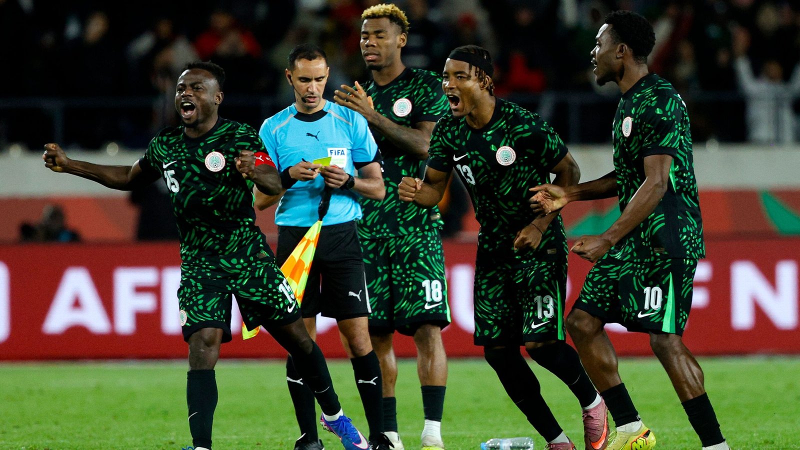 Nigerian players celebrate passionately while facing the referee during an AFCON match, wearing vibrant green jerseys with unique patterns.