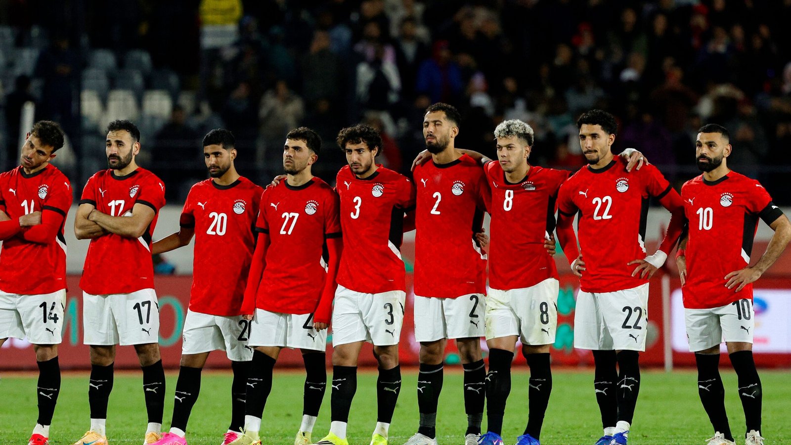 Egyptian national team lined up in red kits, showing unity and focus before a match, with players numbering 14, 17, 20, 3, 2, 8, 22, and 10 prominently displayed.