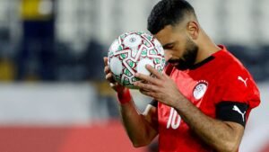 Mohamed Salah, wearing Egypt's red kit, holds a decorated football close to his face in a moment of contemplation on the pitch.