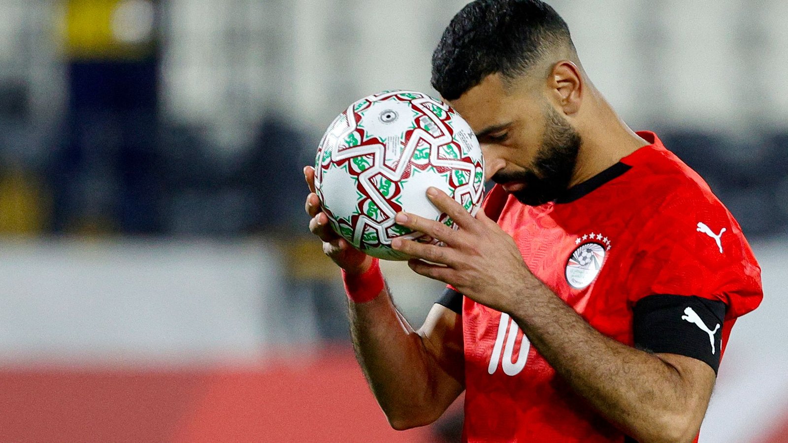 Mohamed Salah, wearing Egypt's red kit, holds a decorated football close to his face in a moment of contemplation on the pitch.