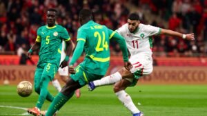 Hakim Ziyech of Morocco, wearing a white kit with green accents, executes a decisive shot as Senegal's defenders, wearing green, close in.