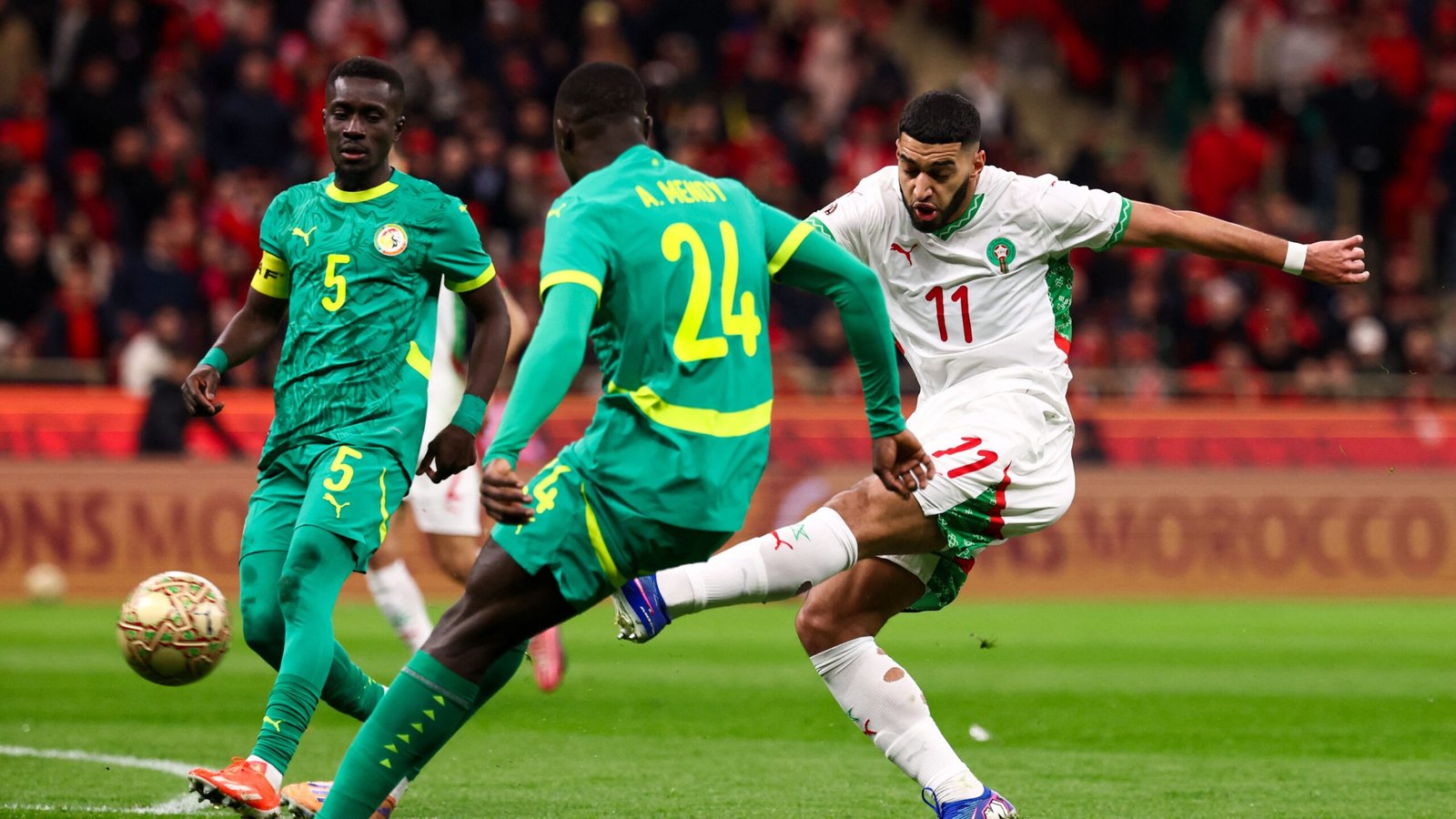 Hakim Ziyech of Morocco, wearing a white kit with green accents, executes a decisive shot as Senegal's defenders, wearing green, close in.