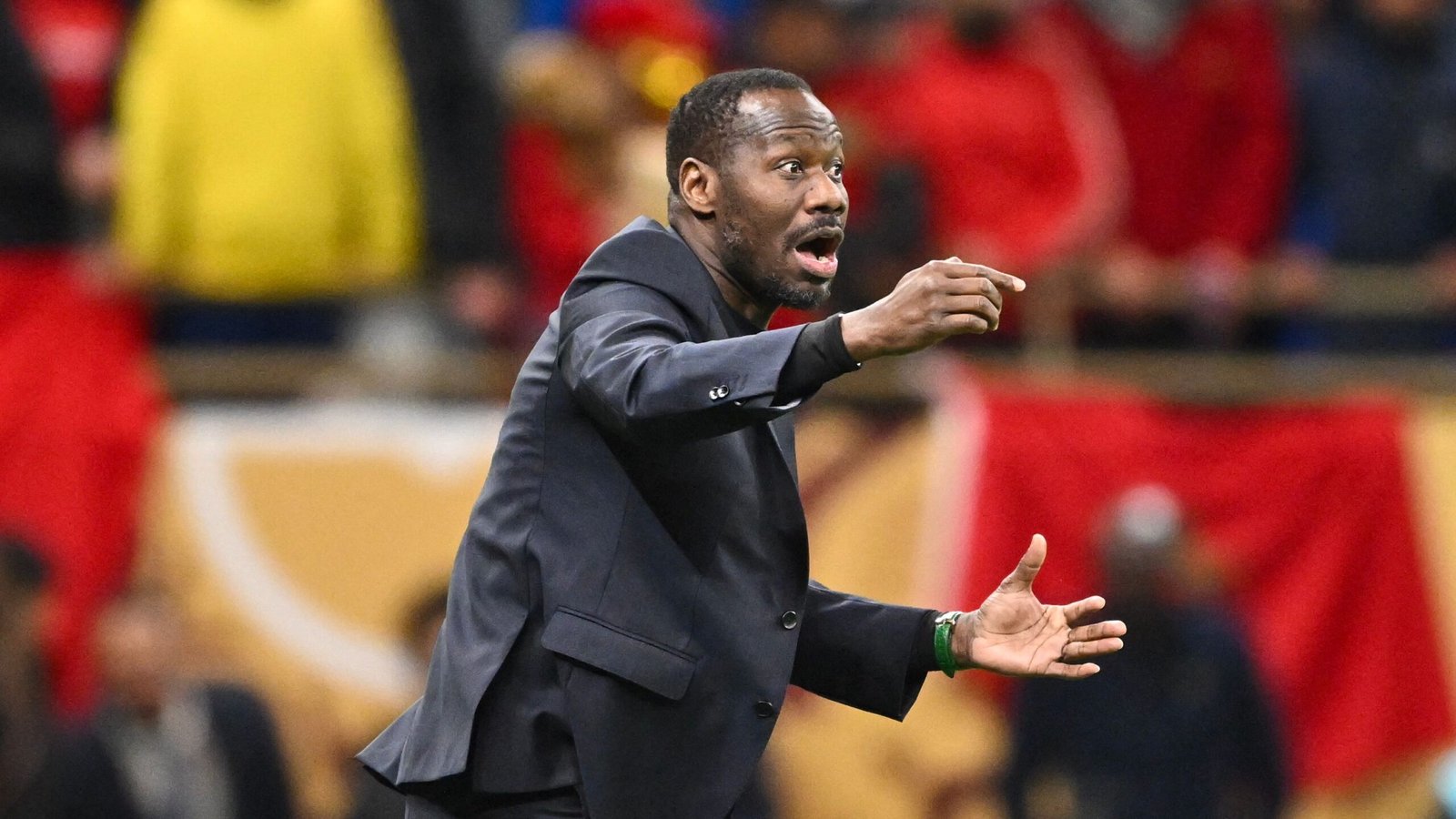 Coach passionately directing players during a match, dressed in a black suit against a backdrop of enthusiastic fans.
