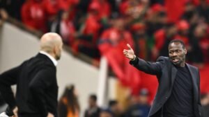 Twee voetbalcoaches in gesprek aan de zijlijn, waarbij één gepassioneerd gebaren maakt. Marokkaanse fans in rode kleding vullen de achtergrond en creëren een elektriserende sfeer.